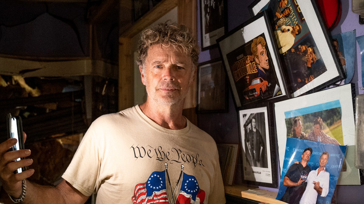 John Schneider standing inside a damaged studio with debris and exposed insulation in the background.