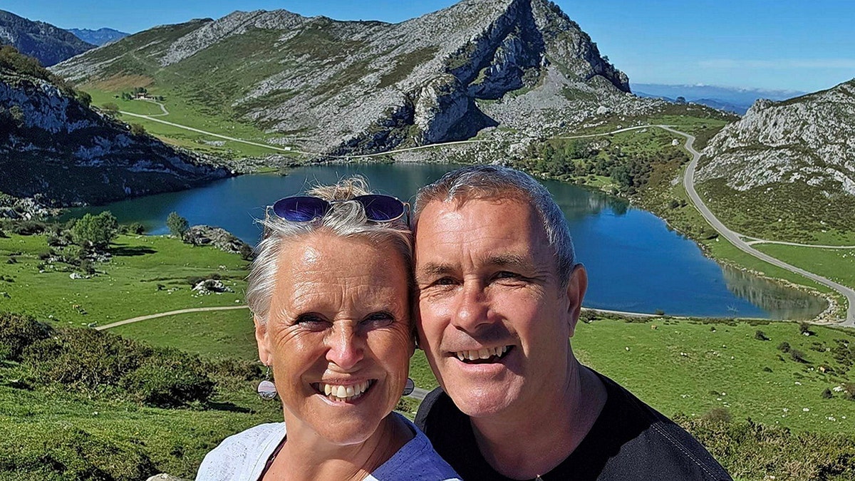 Karen and Myles Davies taking a selfie with a mountain lake and winding road in a scenic green valley.