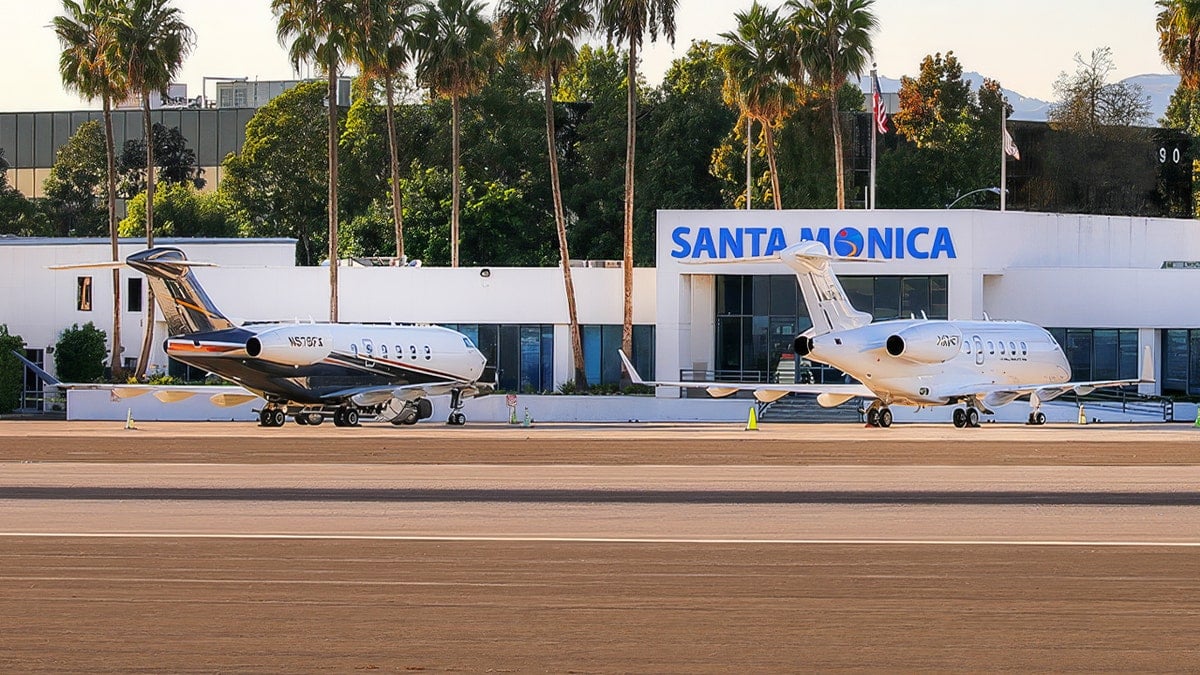 Jets parked on the runway at Santa Monica Airport with palm trees and a small plane taking off in the background.