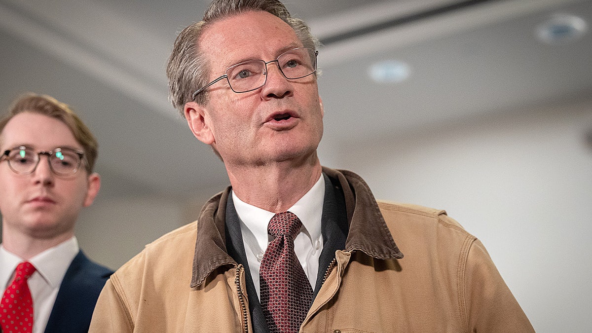 Tim Burchett speaking to a group of reporters while holding a smartphone in a hallway.