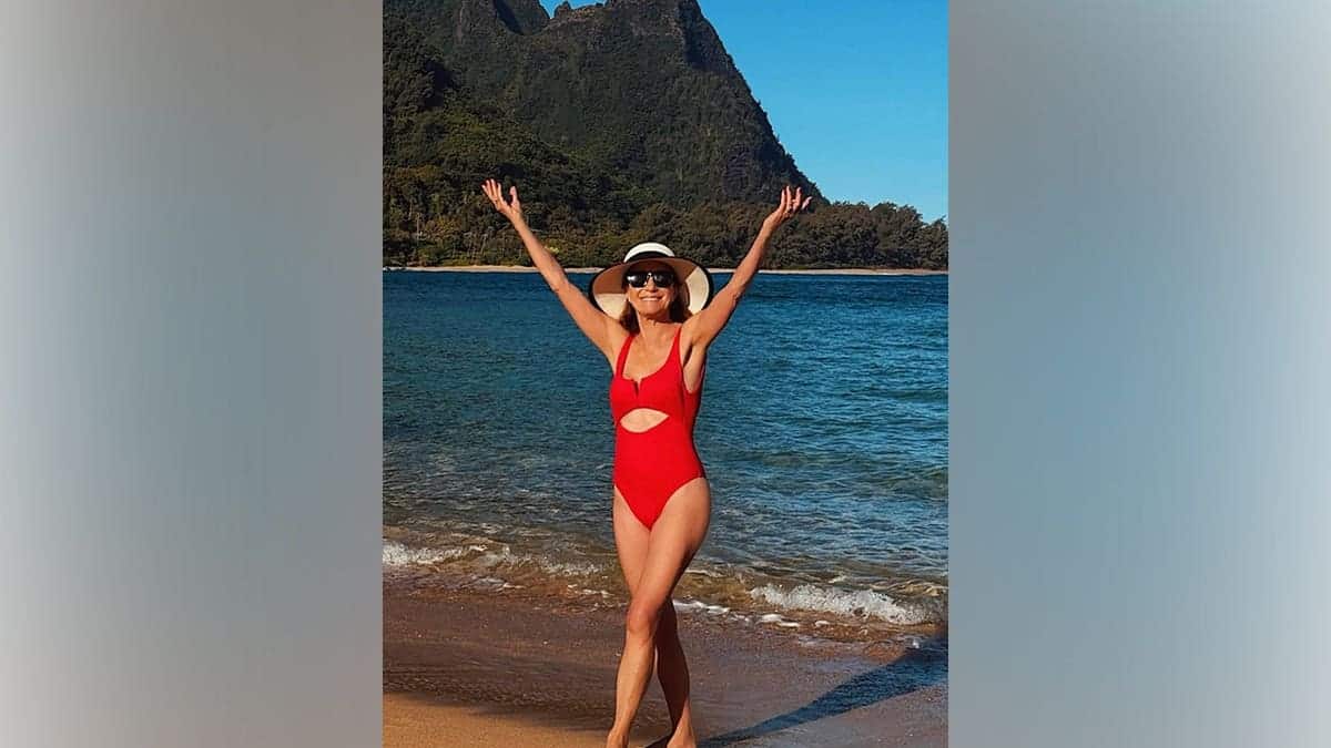 Jane Seymour wearing a red one-piece swimsuit and sun hat walking on a beach with mountains and ocean in the background