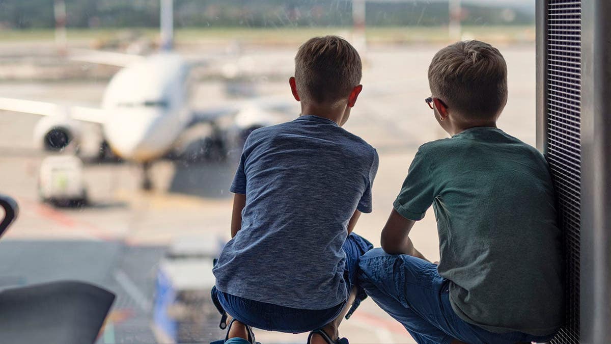 Two young boys crouching by an airport window watching a plane on the tarmac