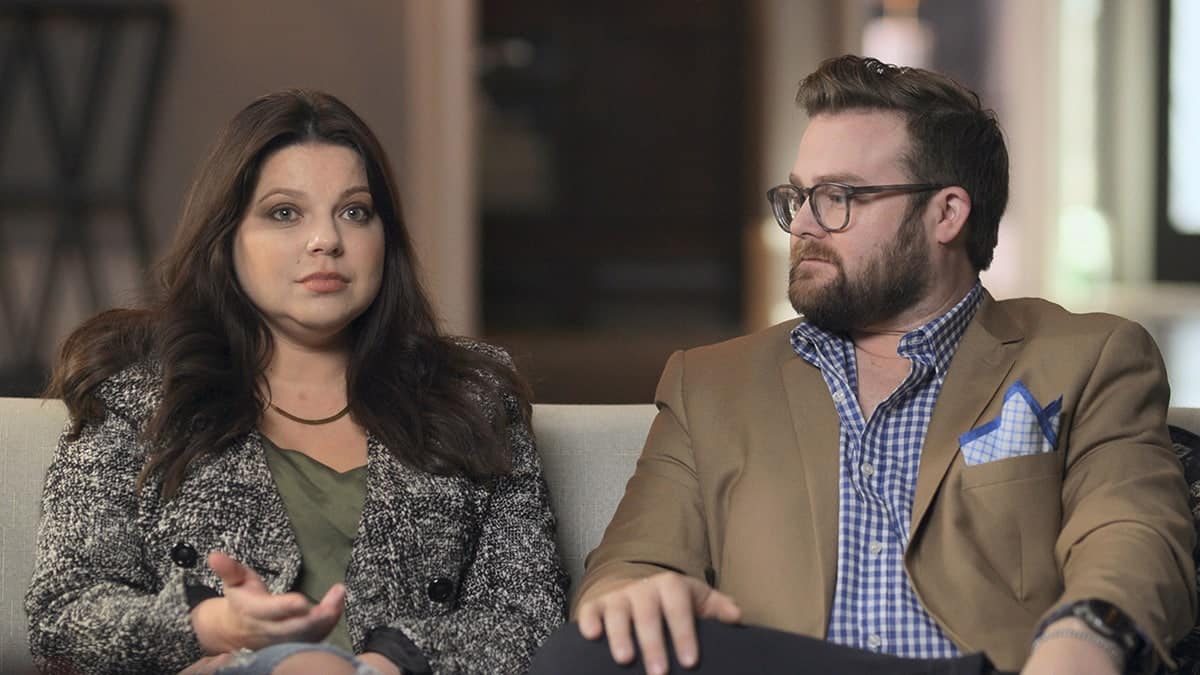 Amy Duggar looking serious sitting on a couch as her husband looks at her.