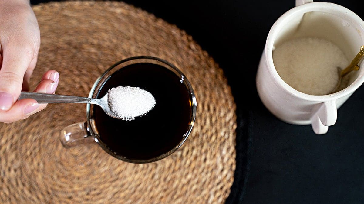 Person pouring sweetener in black coffee seen from overhead angle.