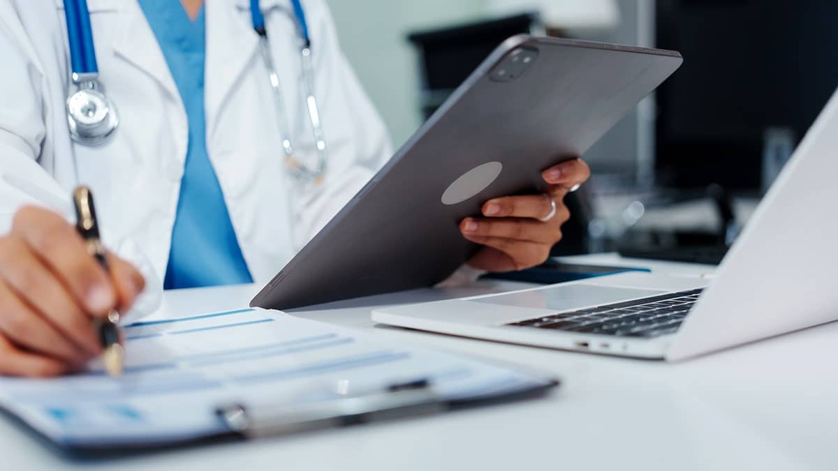 A doctor looks at a tablet and writes on a clipboard.