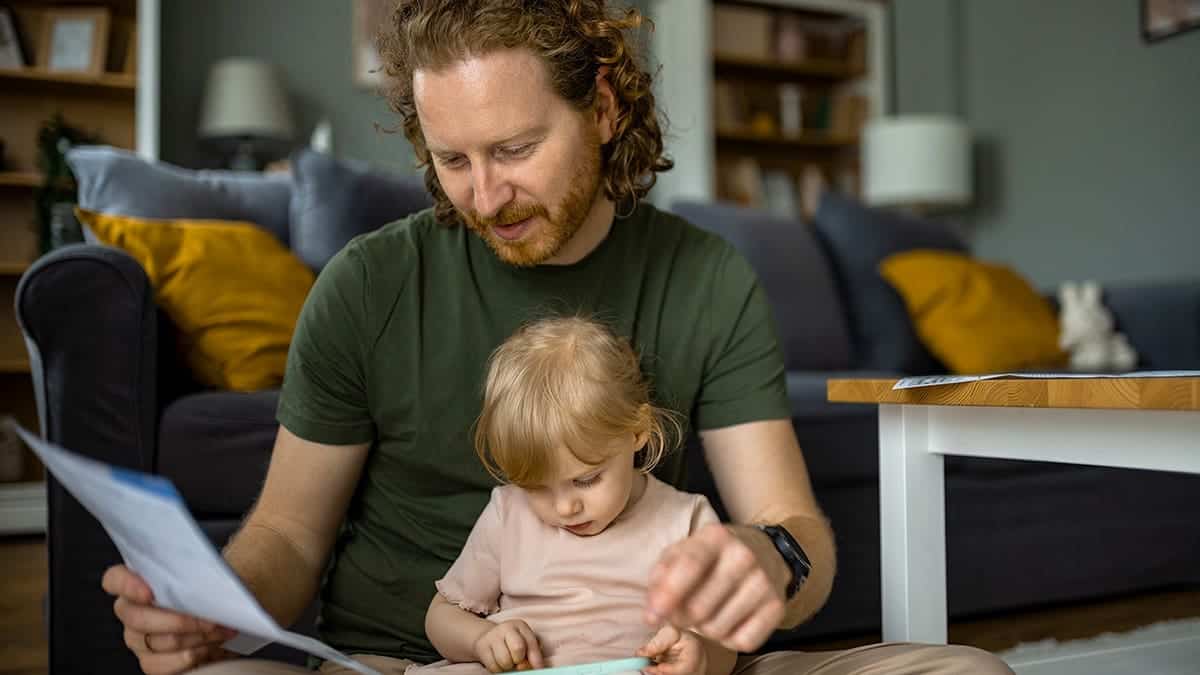 Dad holding baby girl while sorting through bills at home