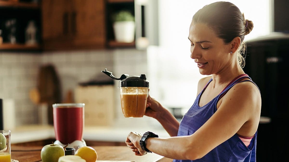Smiling athletic woman drinking a healthy smoothie and checking her wristwatch in a kitchen