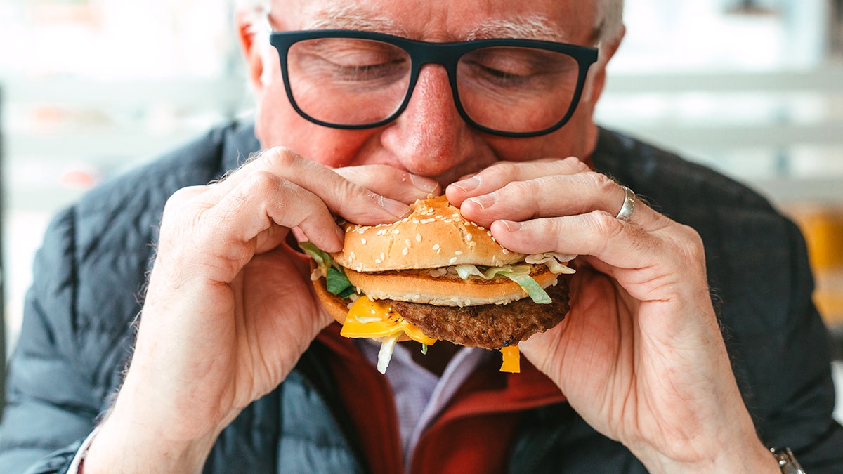 An old man eating a burger indoors.