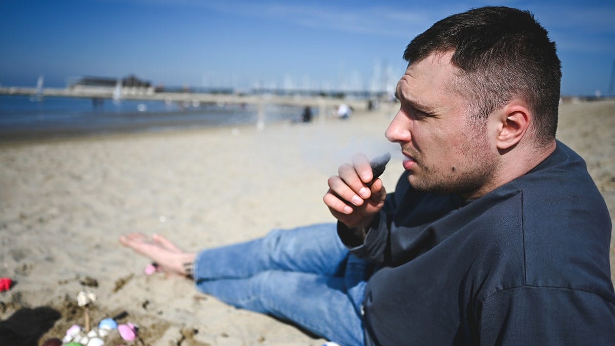 A young man vaping while sitting on a sandy beach near the ocean and pier.