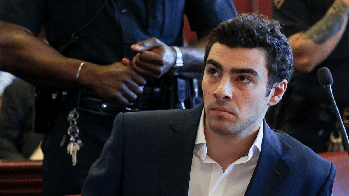 Luigi Mangione in a dark blazer and white shirt with the top two buttons open, leaning to the side in court as an officer stands behind him