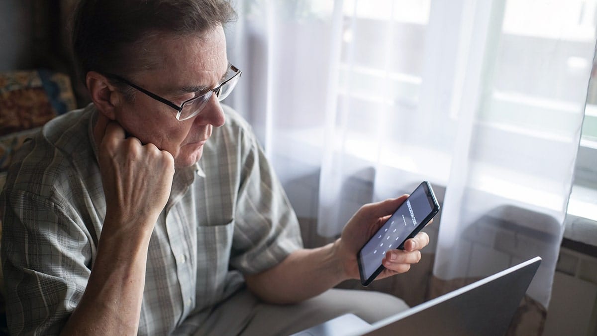A man looks at a security code on his phone with a laptop in front of him.