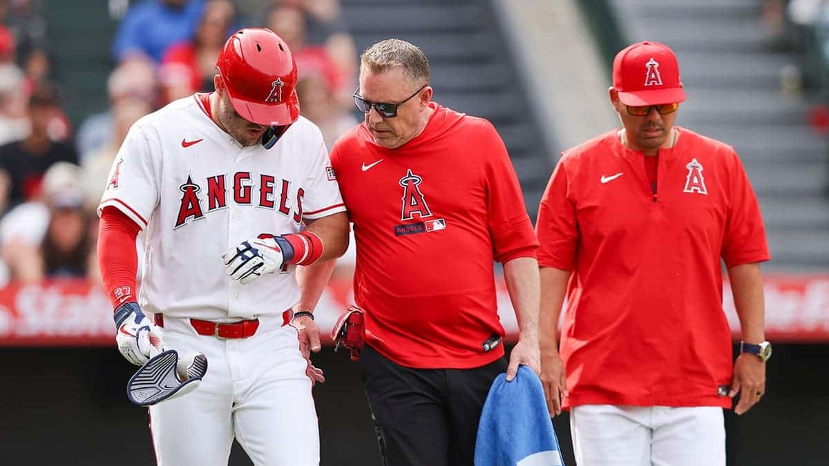 Mike Trout looking down with trainer Mike Frostad and head coach Kurt Suzuki nearby during a baseball game