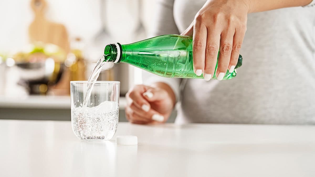 Close up view of female hands pouring tonic water from a green plastic bottle to a glass on kitchen counter.