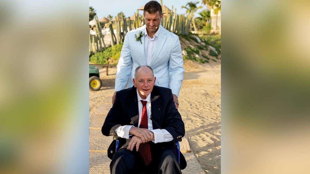 Tim Tebow seen pushing his father in wheelchair at wedding on beach.