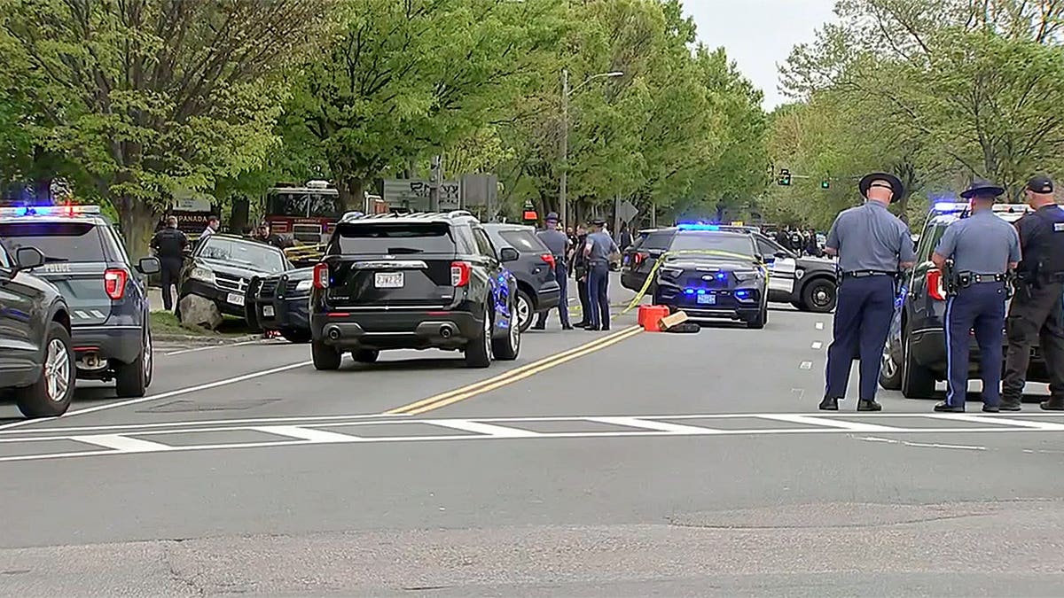 Multiple police vehicles block a roadway in Cambridge, Massachusetts, as officers stand in the street with emergency lights flashing.