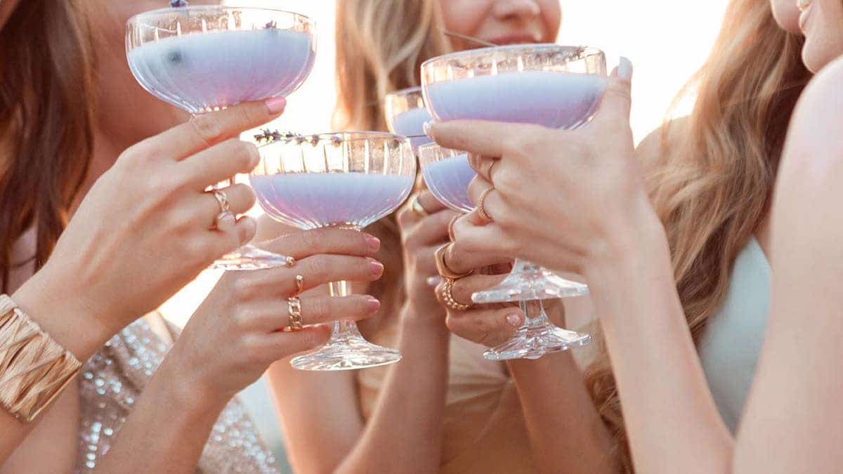 Female friends wearing elegant dresses drinking lavender drinks outdoors at sunset