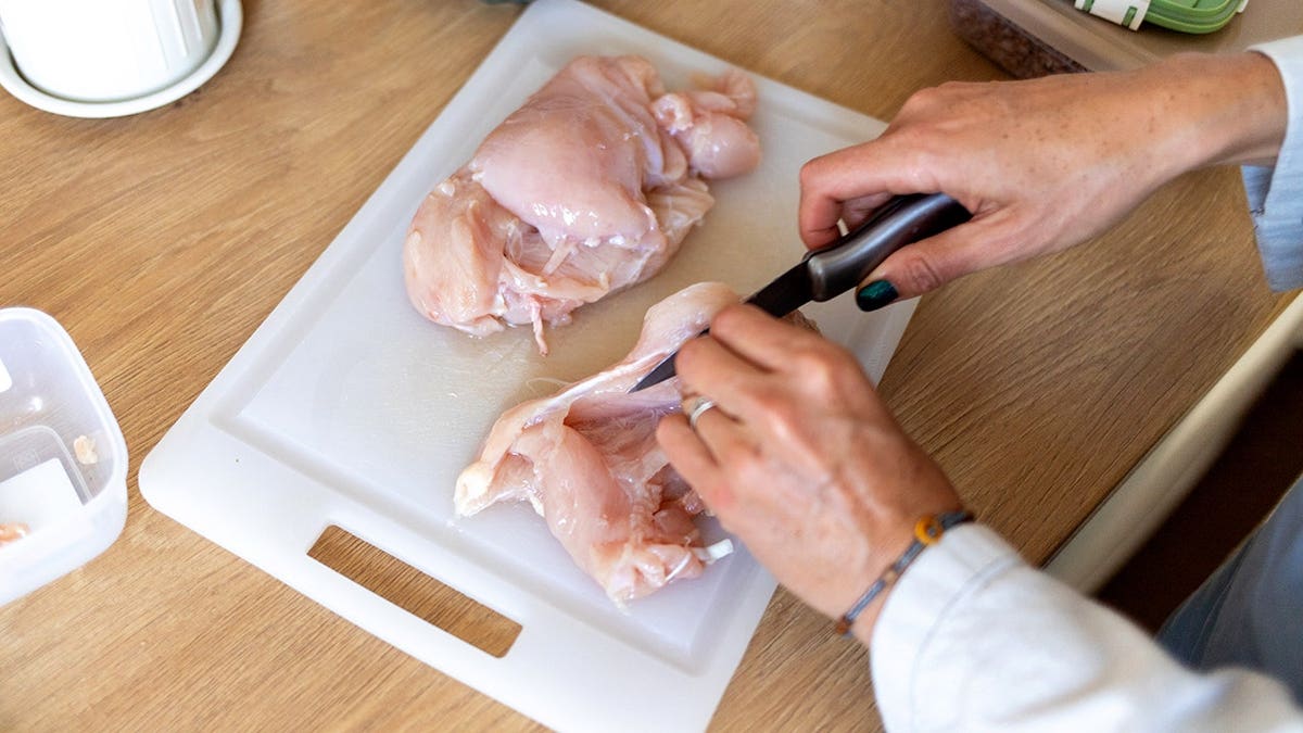 Female hands slicing raw chicken fillet on a cutting board.