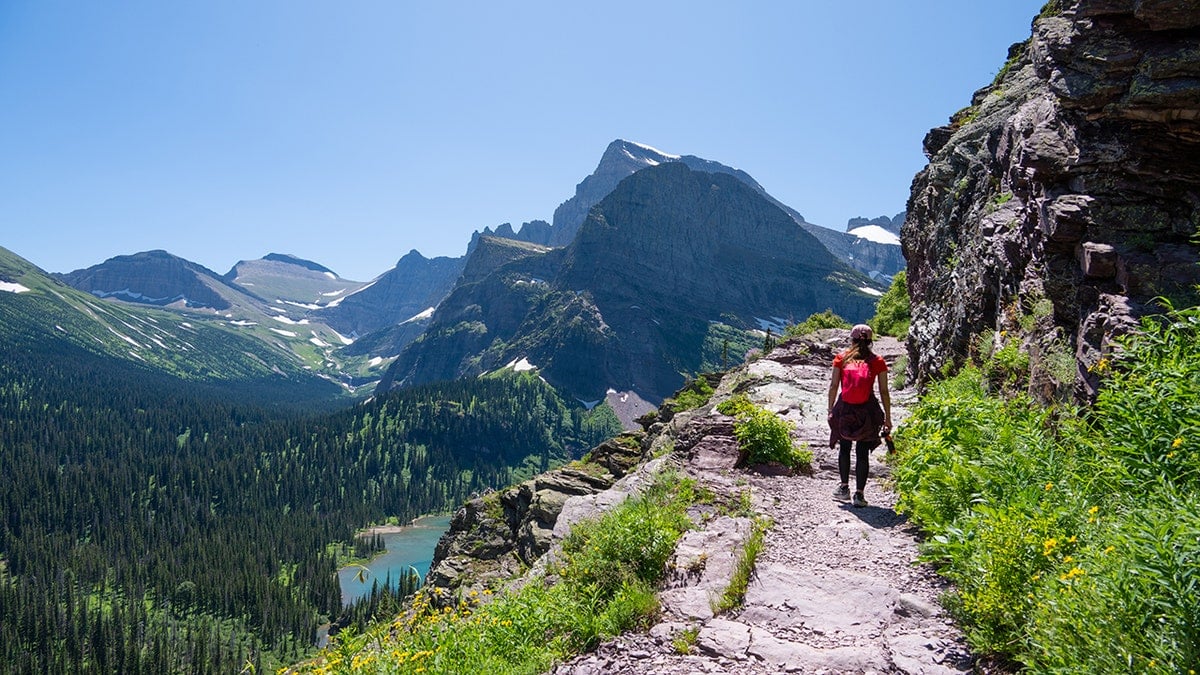 A hiker walking along a narrow mountain trail overlooking an alpine valley in Glacier National Park
