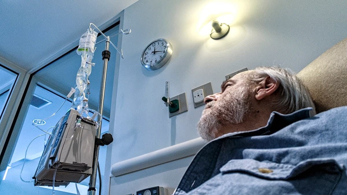 Senior man sitting in a reclining chair looking up at chemotherapy bags hanging from medical equipment.
