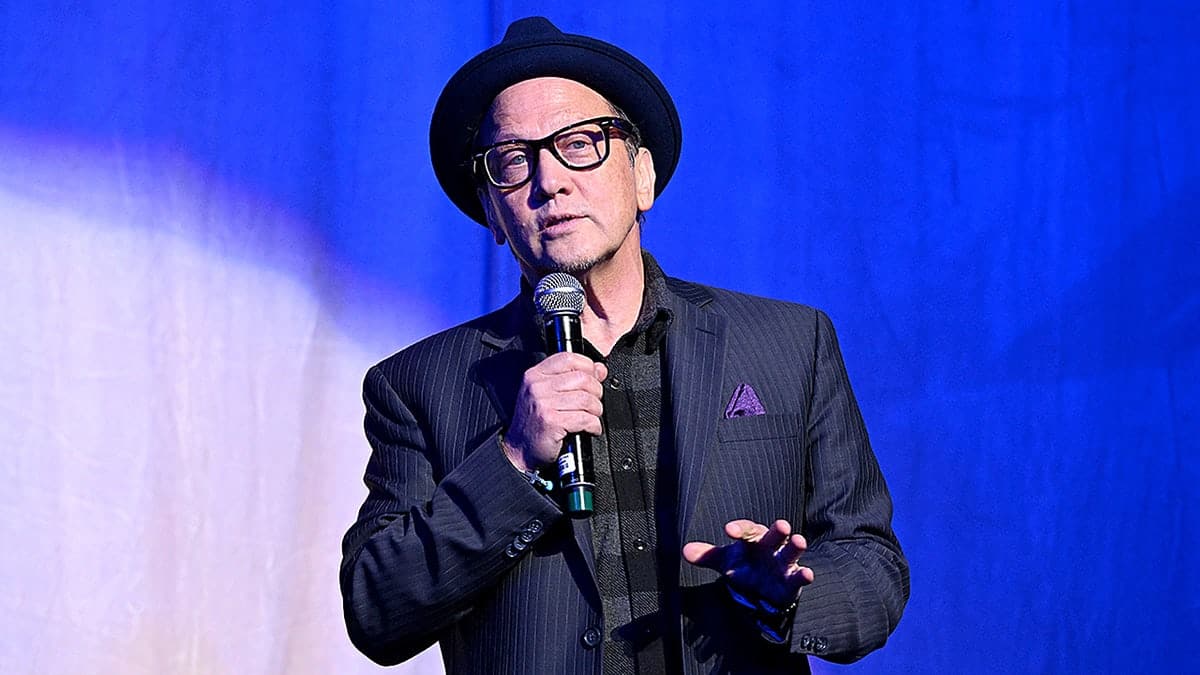 Comedian Rob Schneider holding a microphone on stage at a comedy event