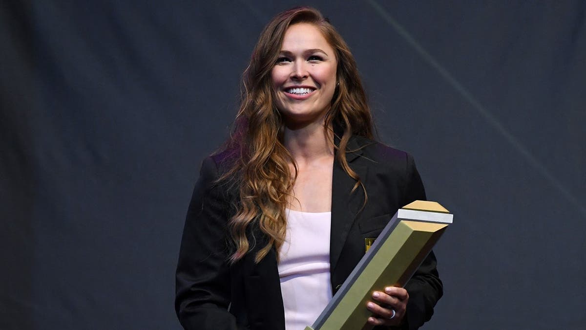Ronda Rousey holding a trophy onstage at the UFC Hall of Fame induction.