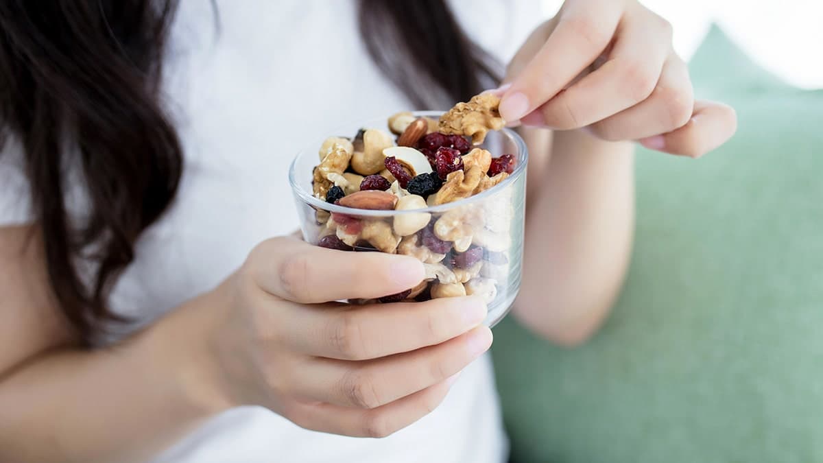 Person holding a glass bowl of mixed nuts and dried fruit, picking up a walnut piece as a healthy snack.