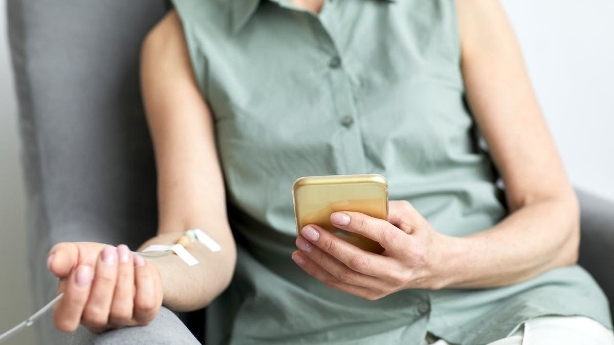 Unrecognizable woman receiving IV drip while using smartphone