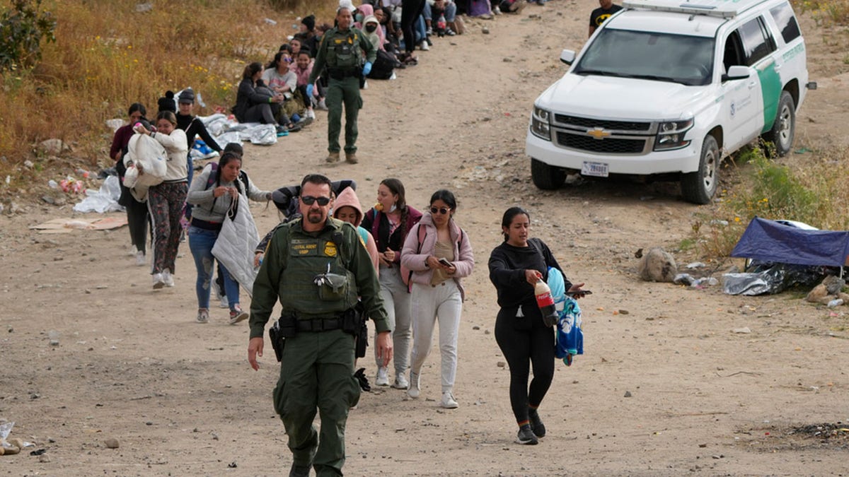 A U.S. Border Patrol agent leading a line of women to a van between two border walls.