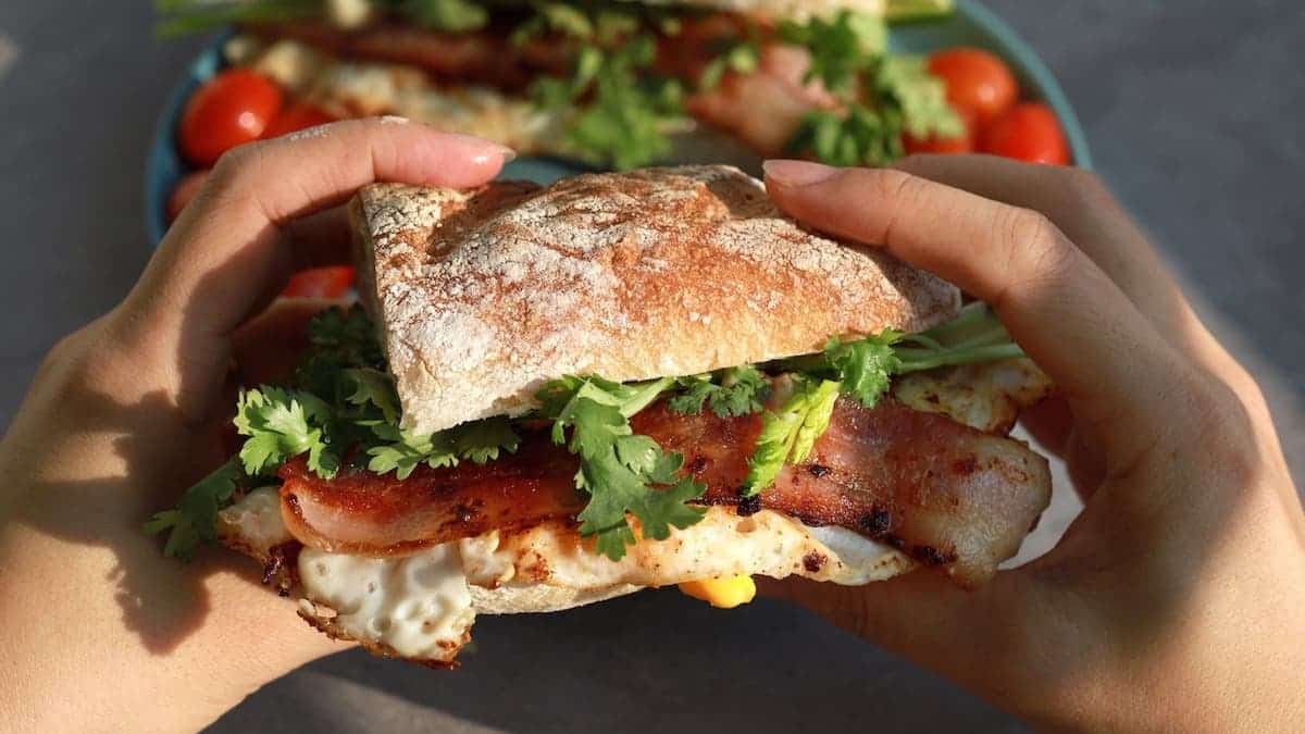 Hand holding a fresh baguette sandwich with bacon, fried egg, parsley, and grilled cherry tomatoes.