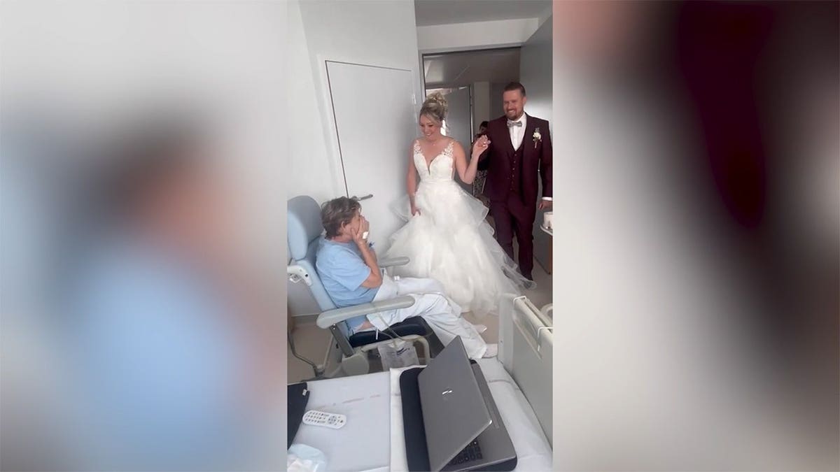 A bride in her wedding gown in a hospital room with the bridal party behind her and her mother in a chair.