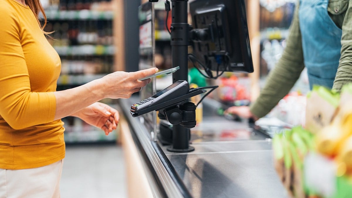 Woman using smartphone to make contactless payment at supermarket checkout.