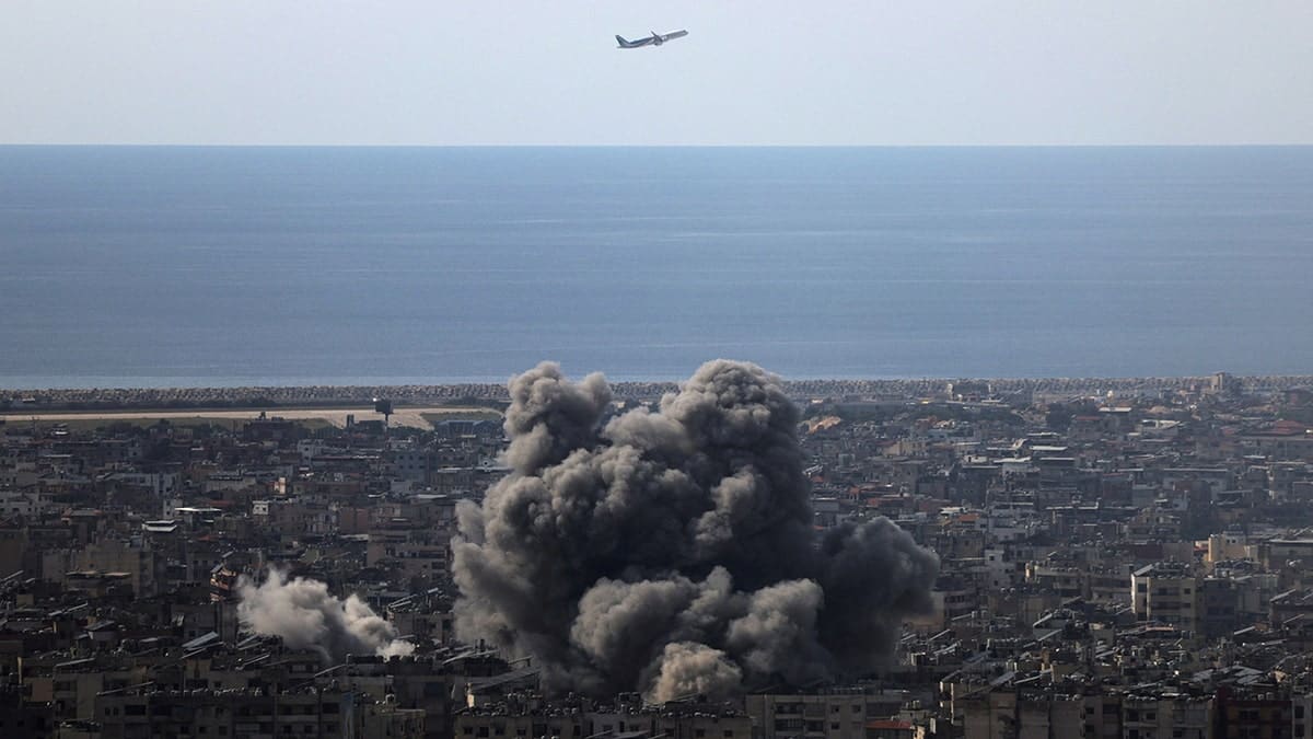 An airplane flies over Beirut's southern suburbs with smoke rising from the ground
