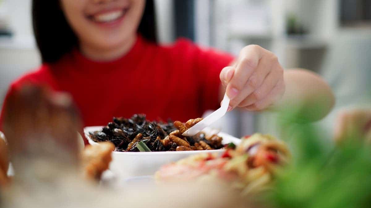 A woman takes a spoonful of bugs from a bowl while smiling.