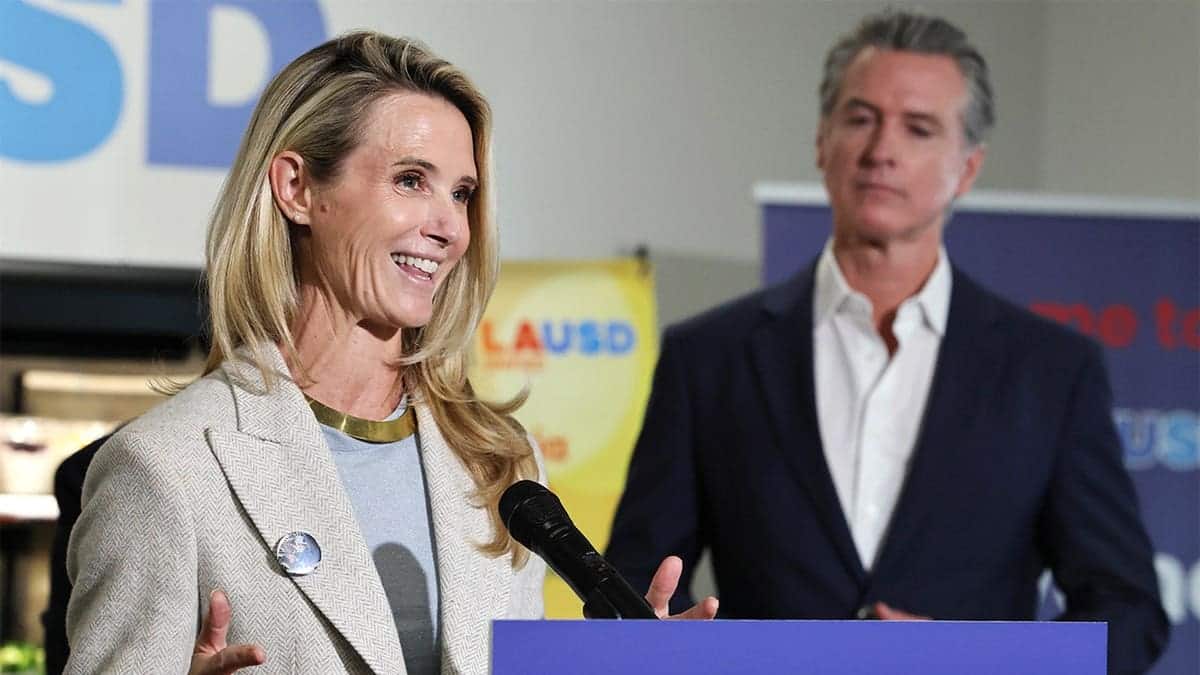 Gavin Newsom looks on as Jennifer Siebel Newsom speaks at a school podium.