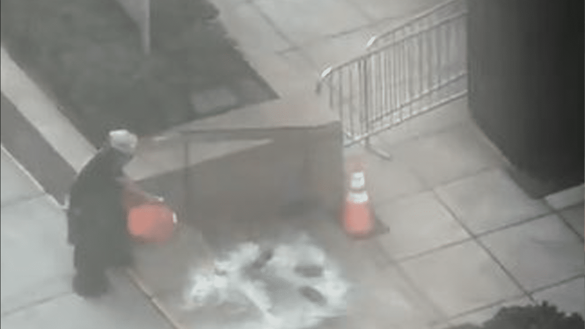 A man in black with a white helmet dumps an orange bucket of gasoline onto the steps outside the building