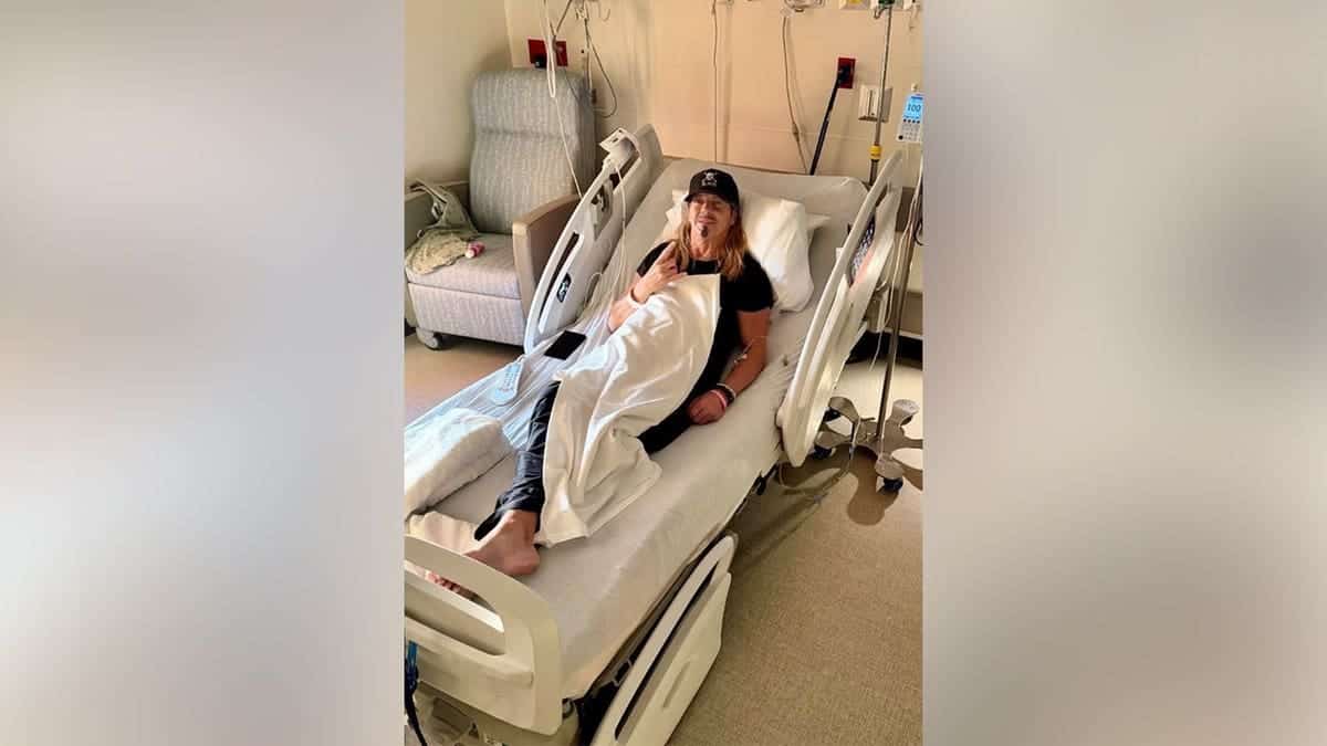 Bret Michaels in a hospital bed in an all-black outfit including a black hat.