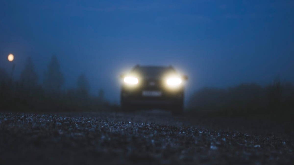 Car with headlights on driving on a road with forest in the background.