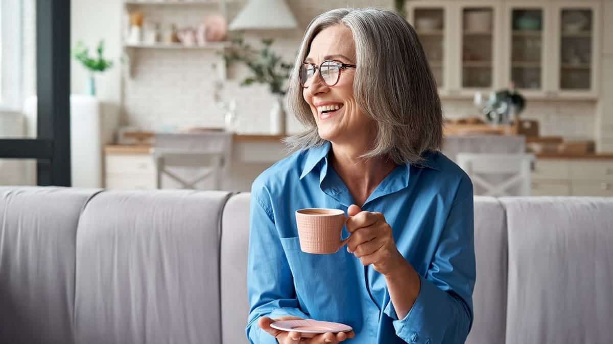 Smiling stylish middle aged 60s lady enjoying resting sitting on couch in modern living room.
