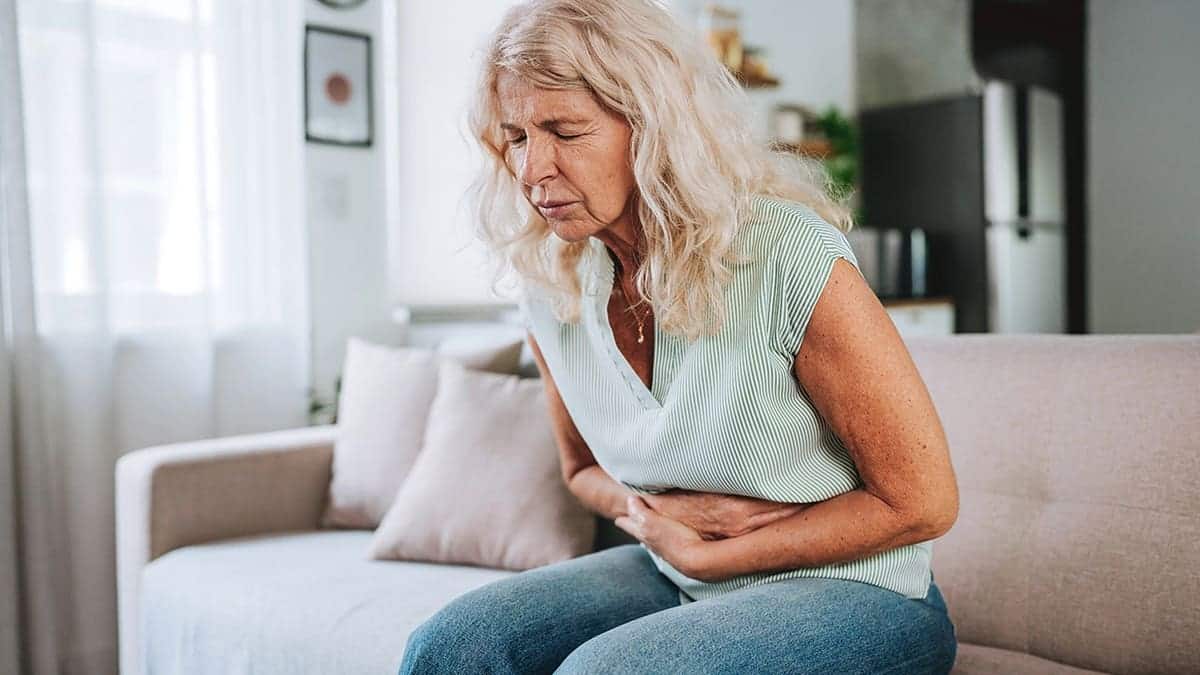 Woman sits on a sofa holding her stomach with a painful expression.