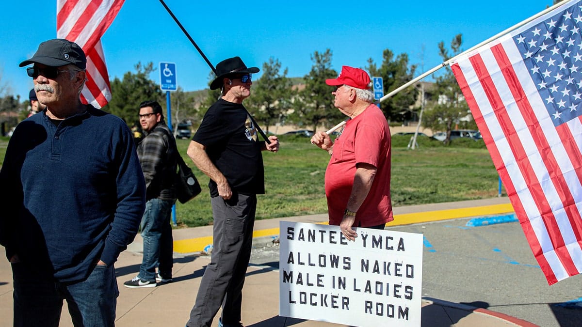 Christynne Wood sitting outdoors as protesters gather with signs in Santee, California.