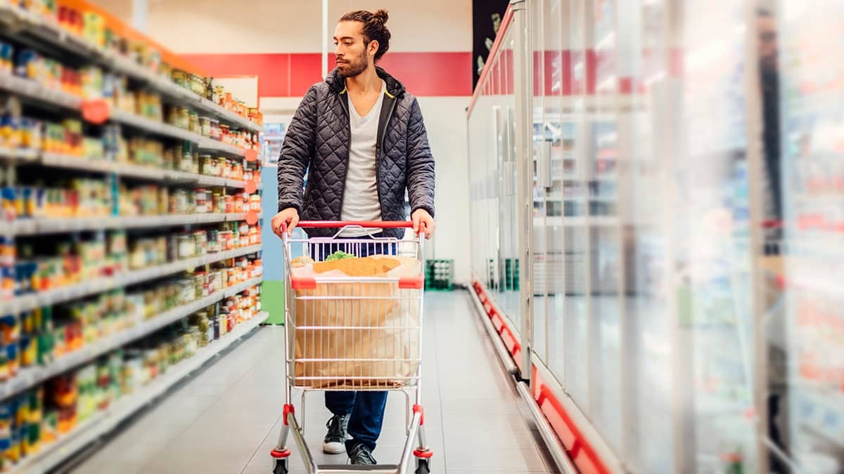 Young man pushing shopping cart through grocery store, looking at canned goods aisle with refrigerators on other side of him.