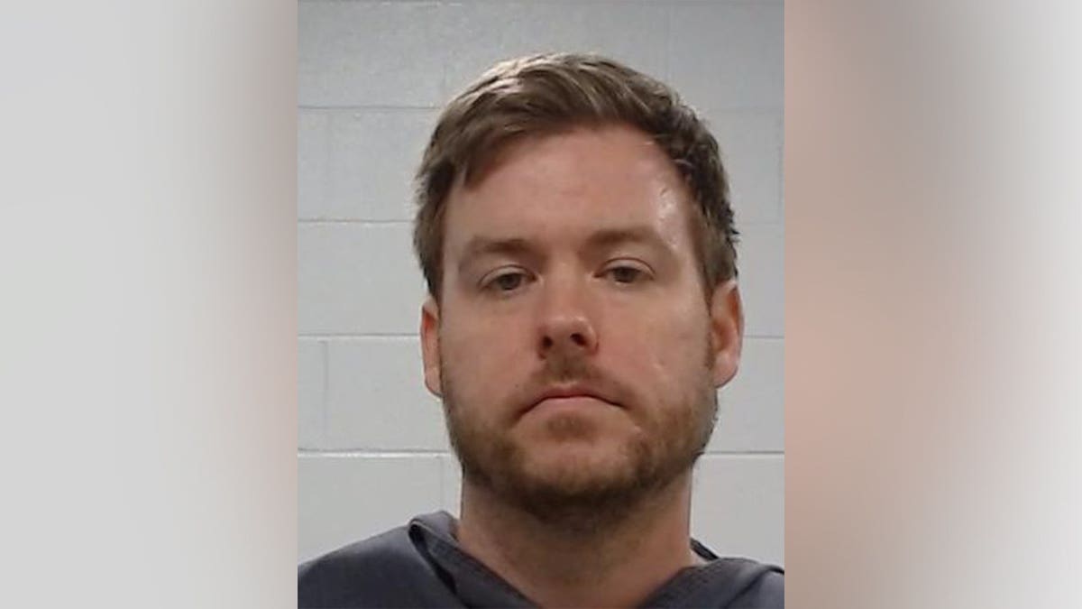 A booking photo of a bearded man in a gray shirt against a cinder-block wall.