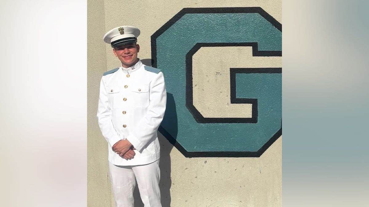 A young man in a white Citadel cadet dress uniform and cap stands smiling beside a large painted