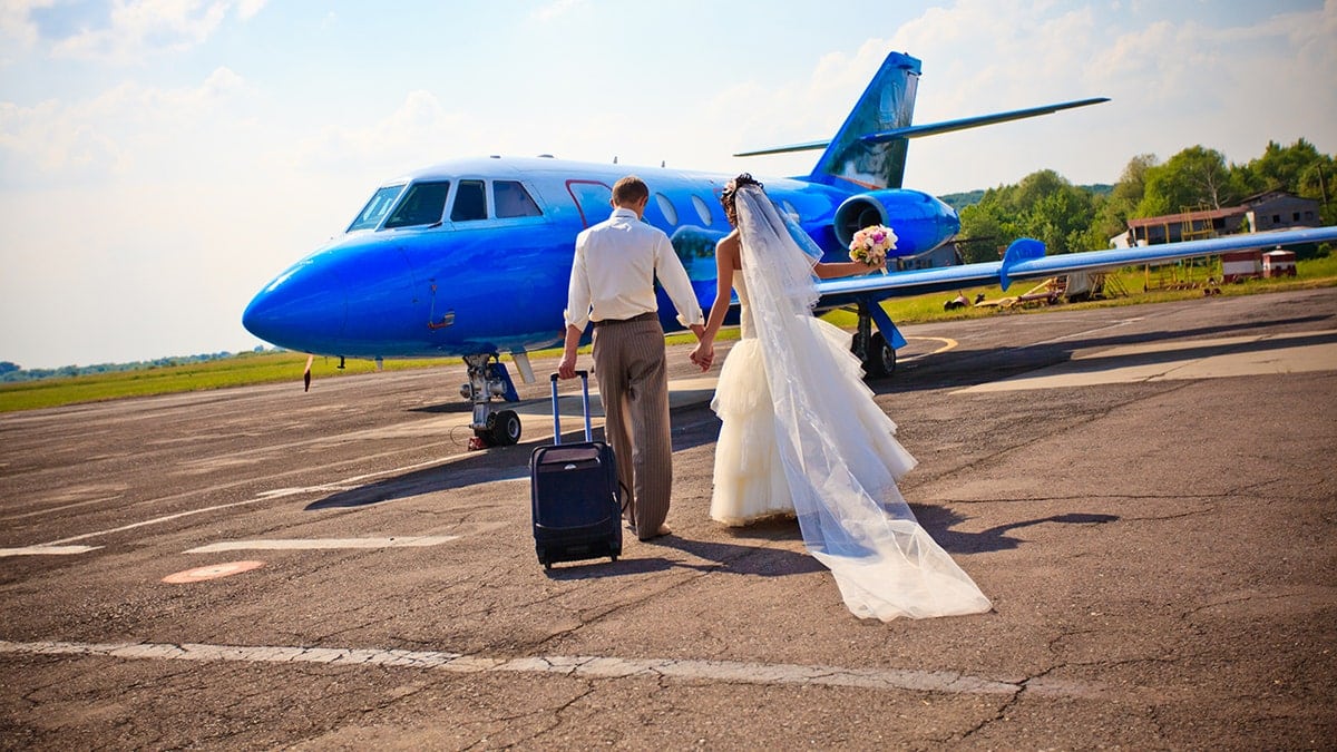 Bride and groom holding hands and walking toward a small blue private jet on an airport runway, beginning their wedding getaway.