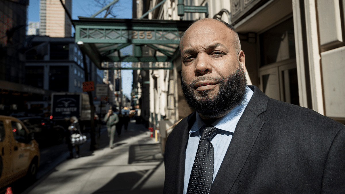 Derrick Parker with a full beard pictured on an NYC sidewalk wearing a dark suit and light blue shirt