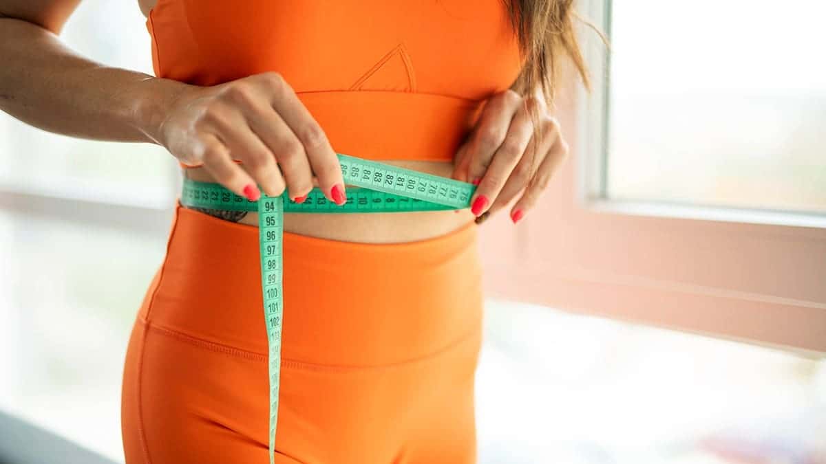 A woman in an orange athletic outfit measuring her waist.