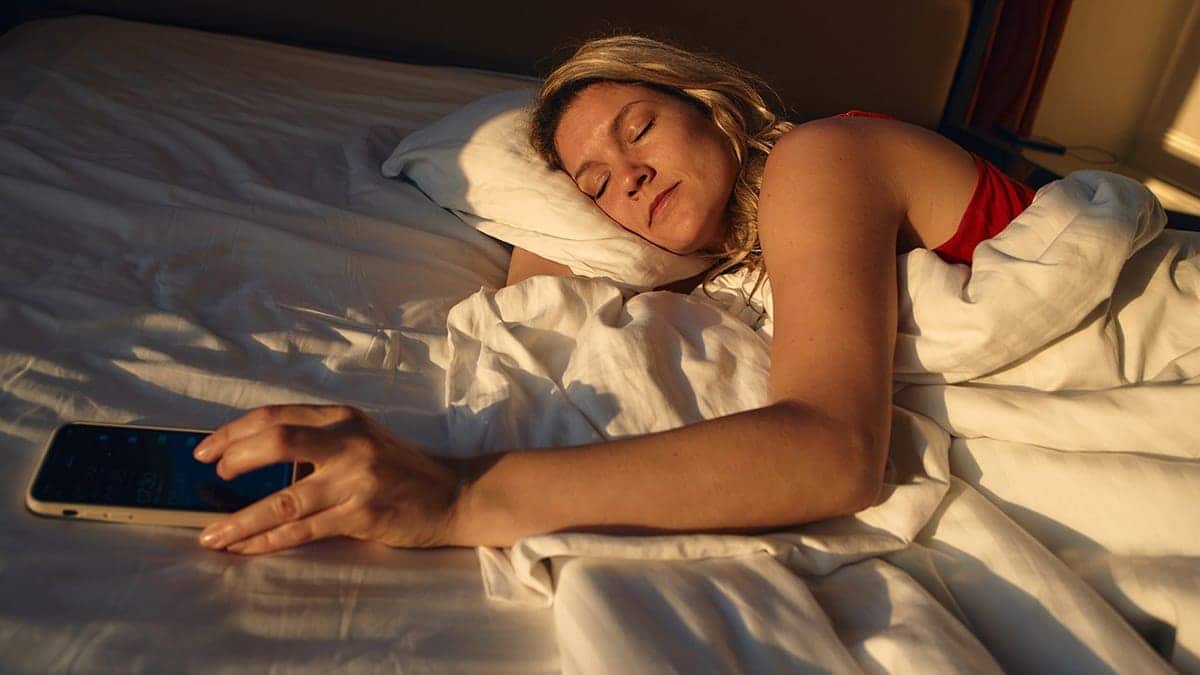 Woman lying in bed reaches for her smartphone.