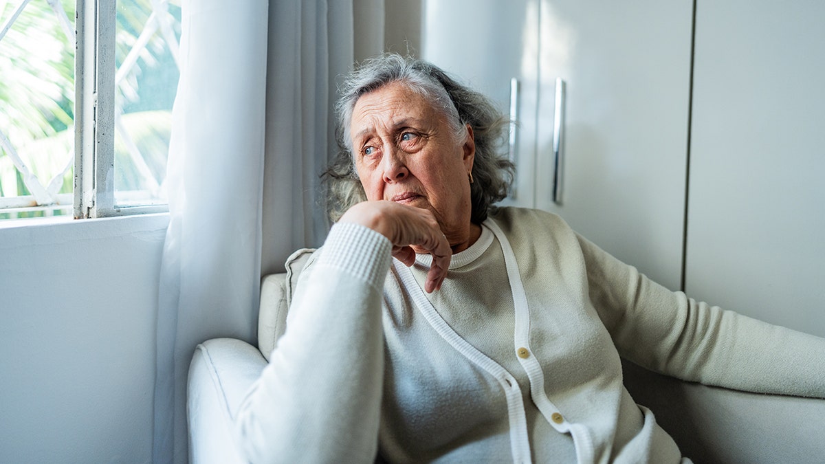 Elderly woman sitting by a window at home, looking outside with a thoughtful expression.