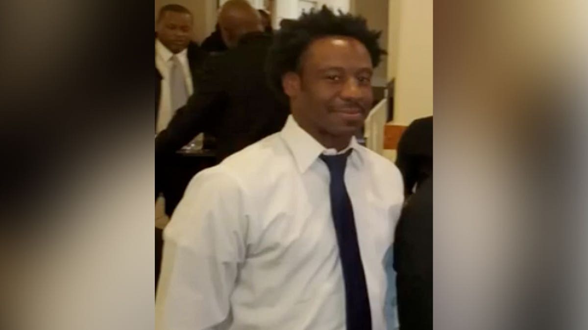 A man in a white dress shirt and dark tie smiles while standing indoors.