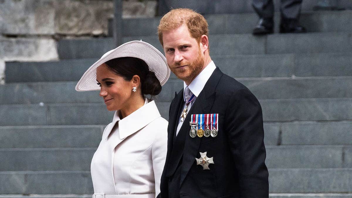 Meghan Markle in a white coat dress and suit walking next to a somber Prince Harry in uniform.